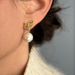 Organic, uneven, and bumpy pearls hang from wing-like element, which is textured, lacy, and studded with diamonds; shown on ear